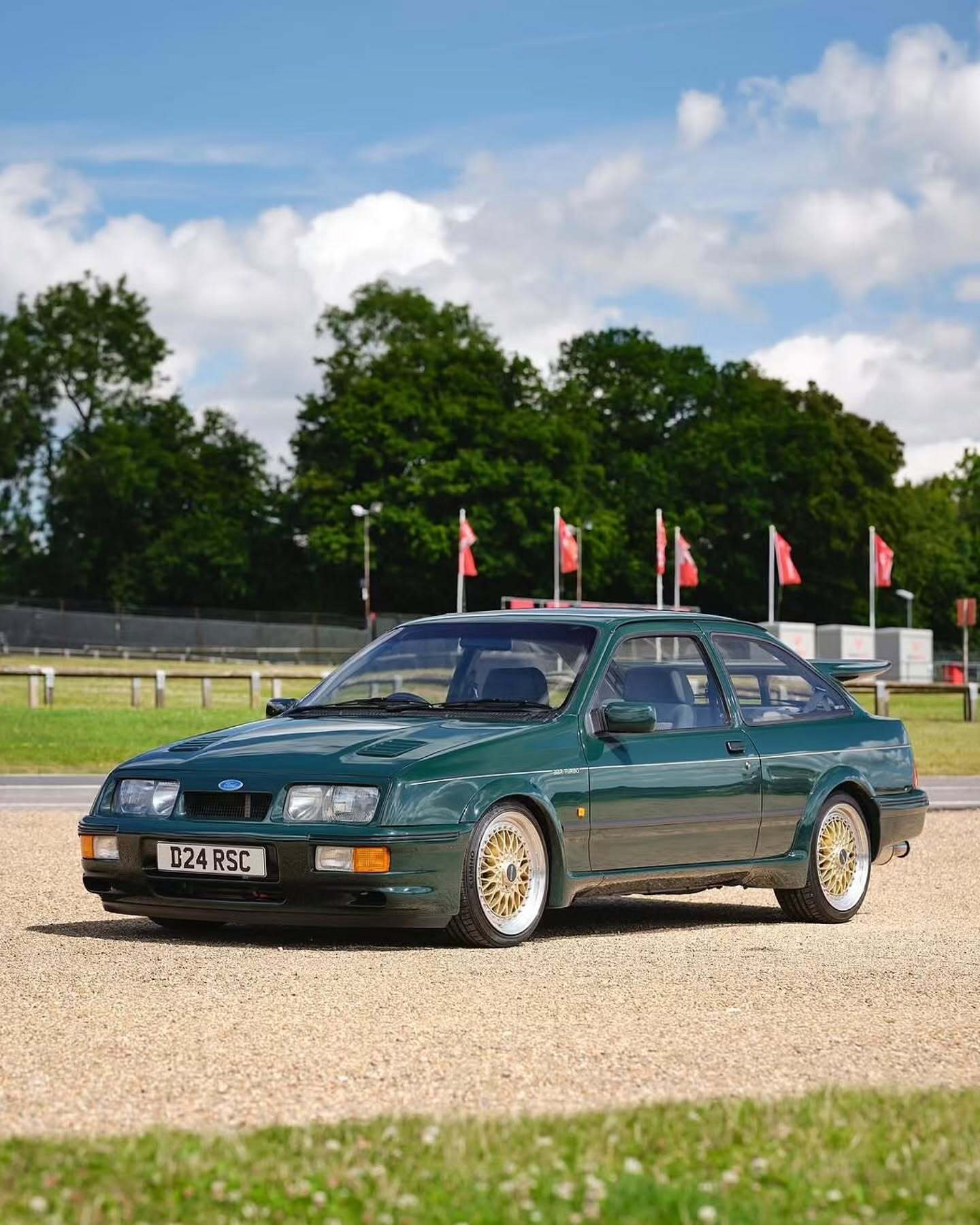 تصویر تمام‌قد و با زاویه باز از خودروی کلاسیک و اسپرت فورد سیرا کاسورث (Ford Sierra Cosworth) با رنگ تیره، که باله عظیم عقب و گلگیرهای برجسته آن در دهه ۱۹۸۰ میلادی آن را به یک نماد سرعت و سلطه بر پیست‌های مسابقه تبدیل کرد.